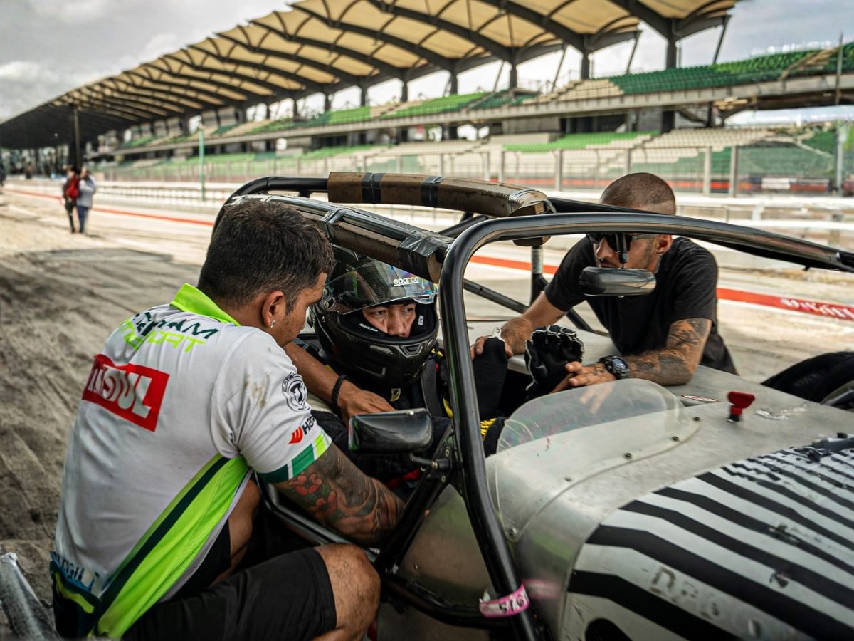Driver strapped into a race car cockpit with two crew members leaning in