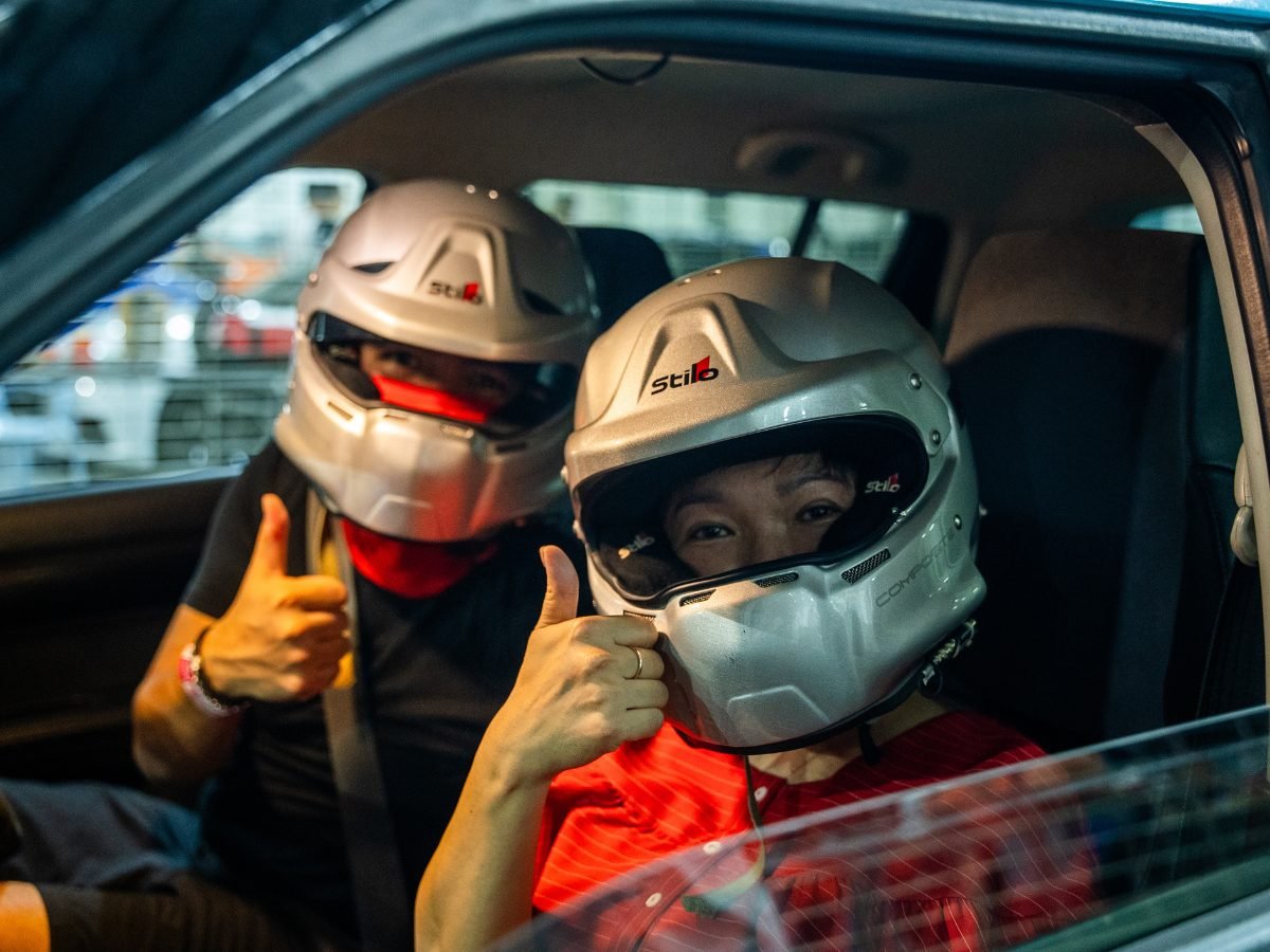 Two people in full-face helmets giving thumbs up from inside a car