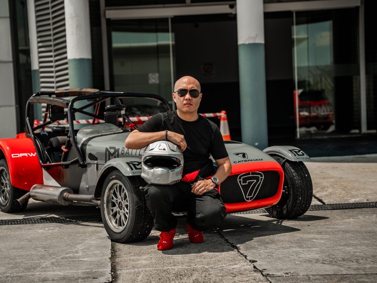 Nicky Lim crouched with a helmet beside a grey and orange Caterham Seven